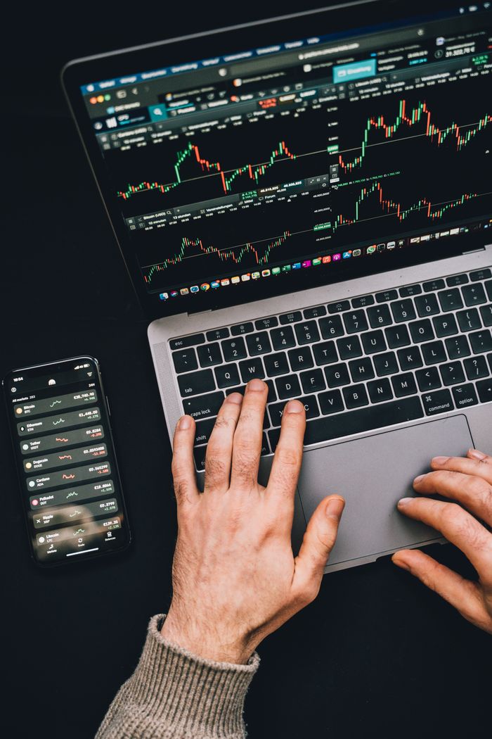 Mains en train de taper sur un ordinateur portable, avec des graphiques de cryptomonnaies et l’écran d’un smartphone visibles, dans un univers centré sur la finance numérique.